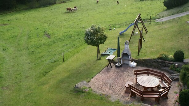 Un paysage rural avec un champ d’herbe, une balançoire en bois et une terrasse en briques avec une table et des bancs.