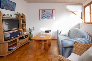 Un salon confortable avec un téléviseur, un meuble de rangement en bois, une petite table, un canapé et une fenêtre avec des stores.