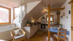 A kitchen with wooden cabinets, a dining table with chairs, a wicker chair with a cushion, and a potted plant on the counter. - Apartment 'Anne's' with Balcony and Wi-Fi (Bad Waldsee)