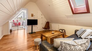 Una habitación estilo loft con techo inclinado, una mesa de comedor de madera, un sofá con cojines estampados, un televisor y una silla roja.