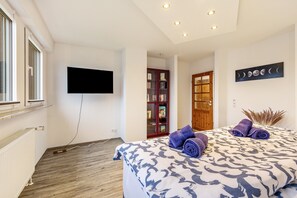 Un dormitorio con una cama, un televisor de pantalla plana, una estantería y una puerta que da a otra habitación.