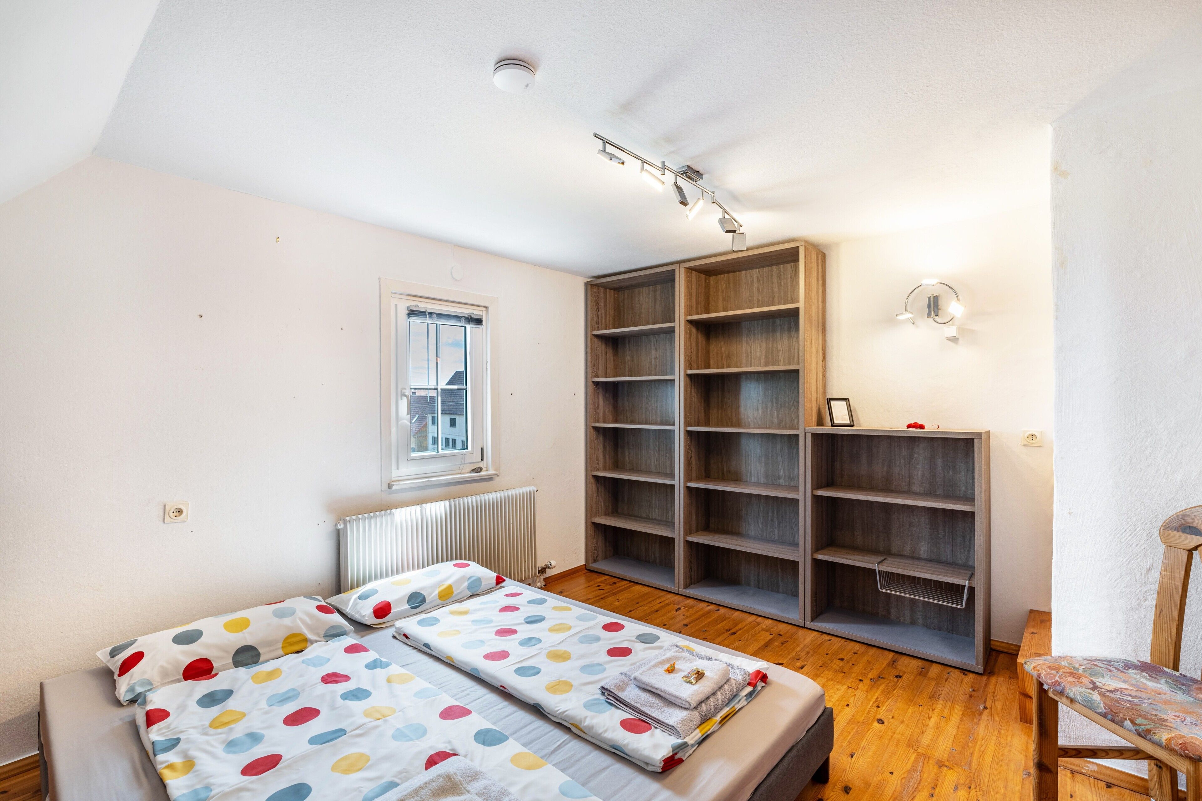 A bedroom with a bed, a window, a radiator, a bookshelf, and a chair.