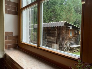 A log cabin with a wheelbarrow outside, viewed through a window with a wooden frame and a brick wall.
