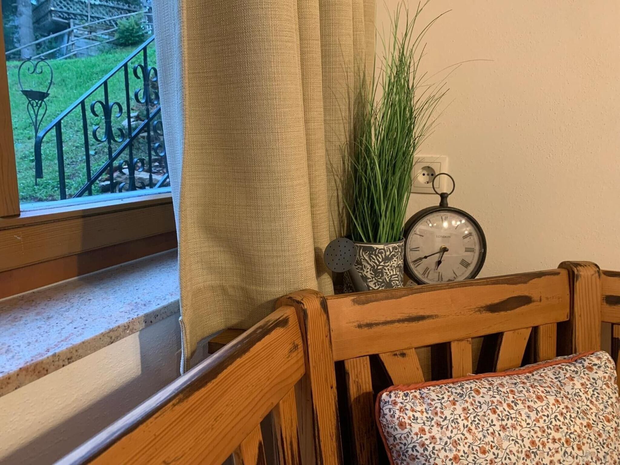 A wooden chair with a floral cushion, a potted plant, and a vintage clock on a windowsill.