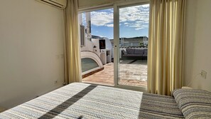 Een slaapkamer met een bed, een schuifdeur die leidt naar een buitenruimte met een terras en uitzicht op een gebouw.