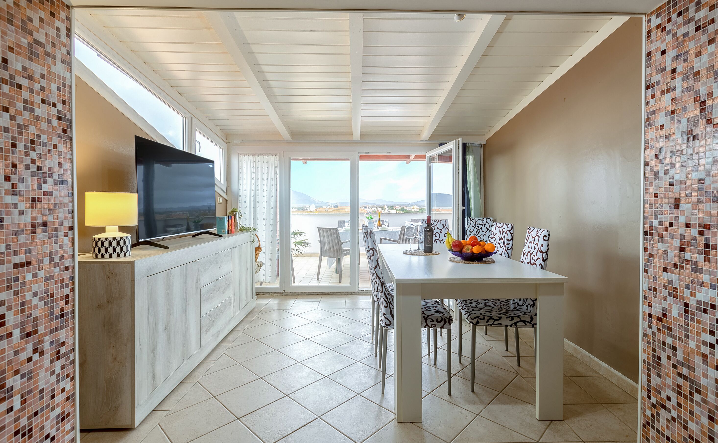 A modern dining area with a TV, dining table, chairs, and a view of the sea.