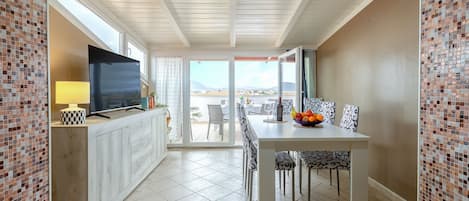 A modern dining area with a TV, dining table, chairs, and a view of the sea.
