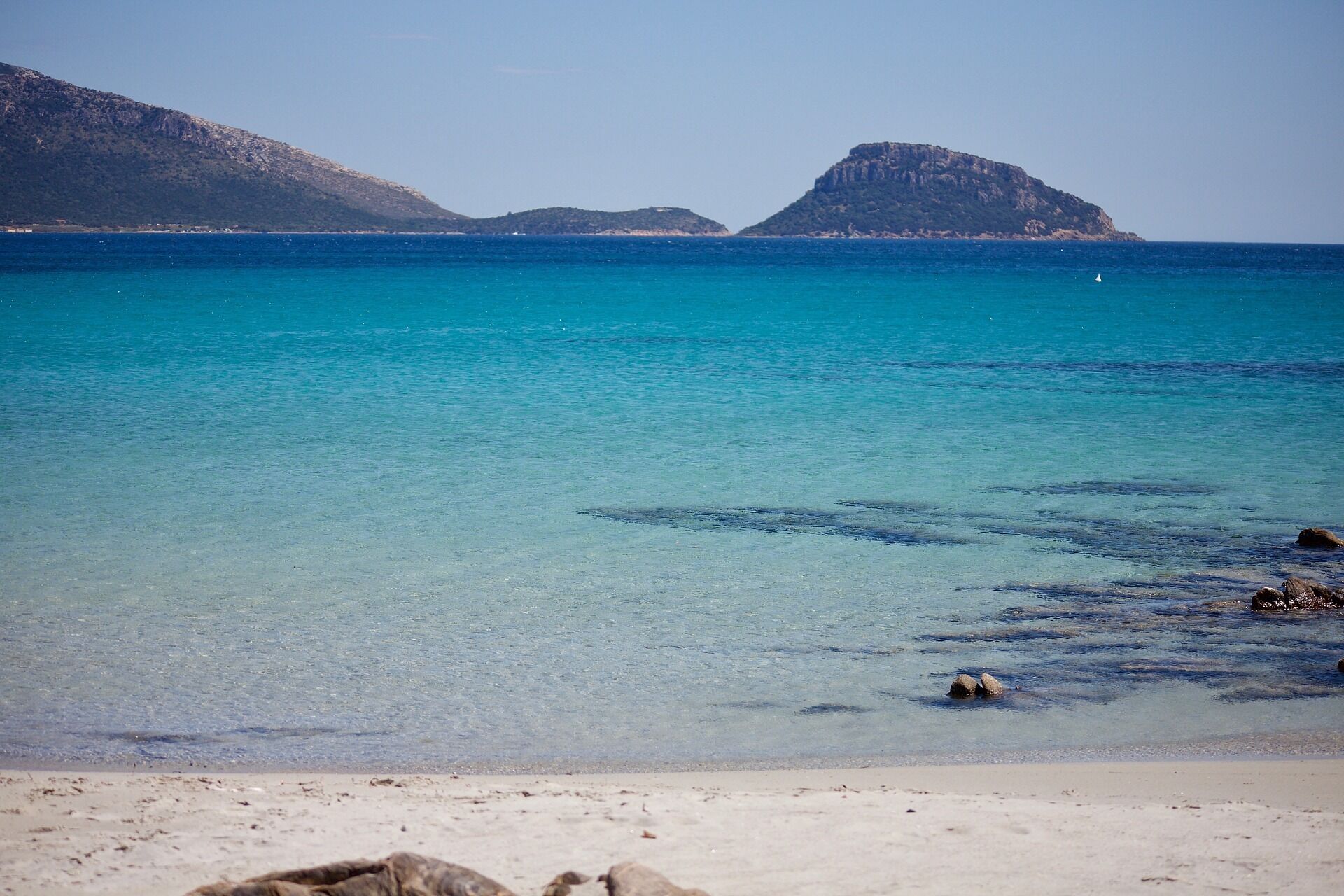 A beach with clear turquoise water, rocky shoreline, and distant mountains.
