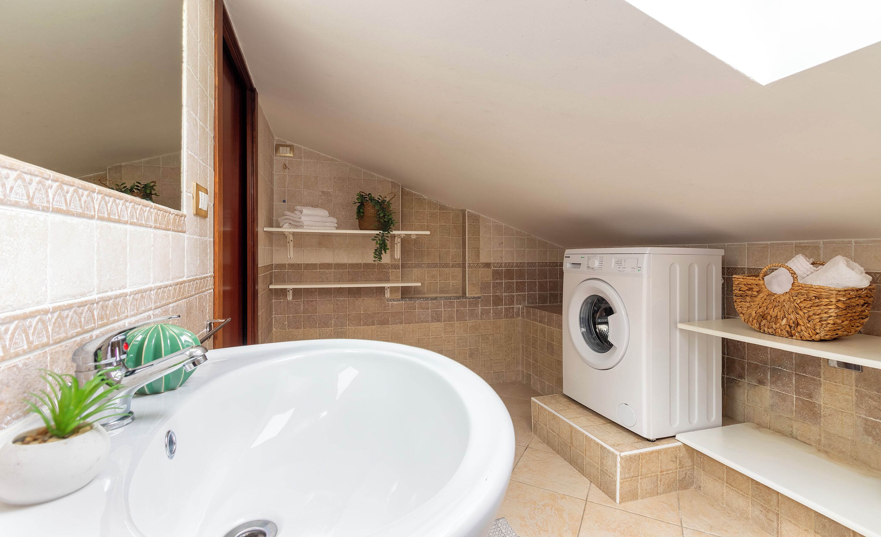A modern bathroom with a large sink, a built-in washing machine, and shelves with towels and plants.