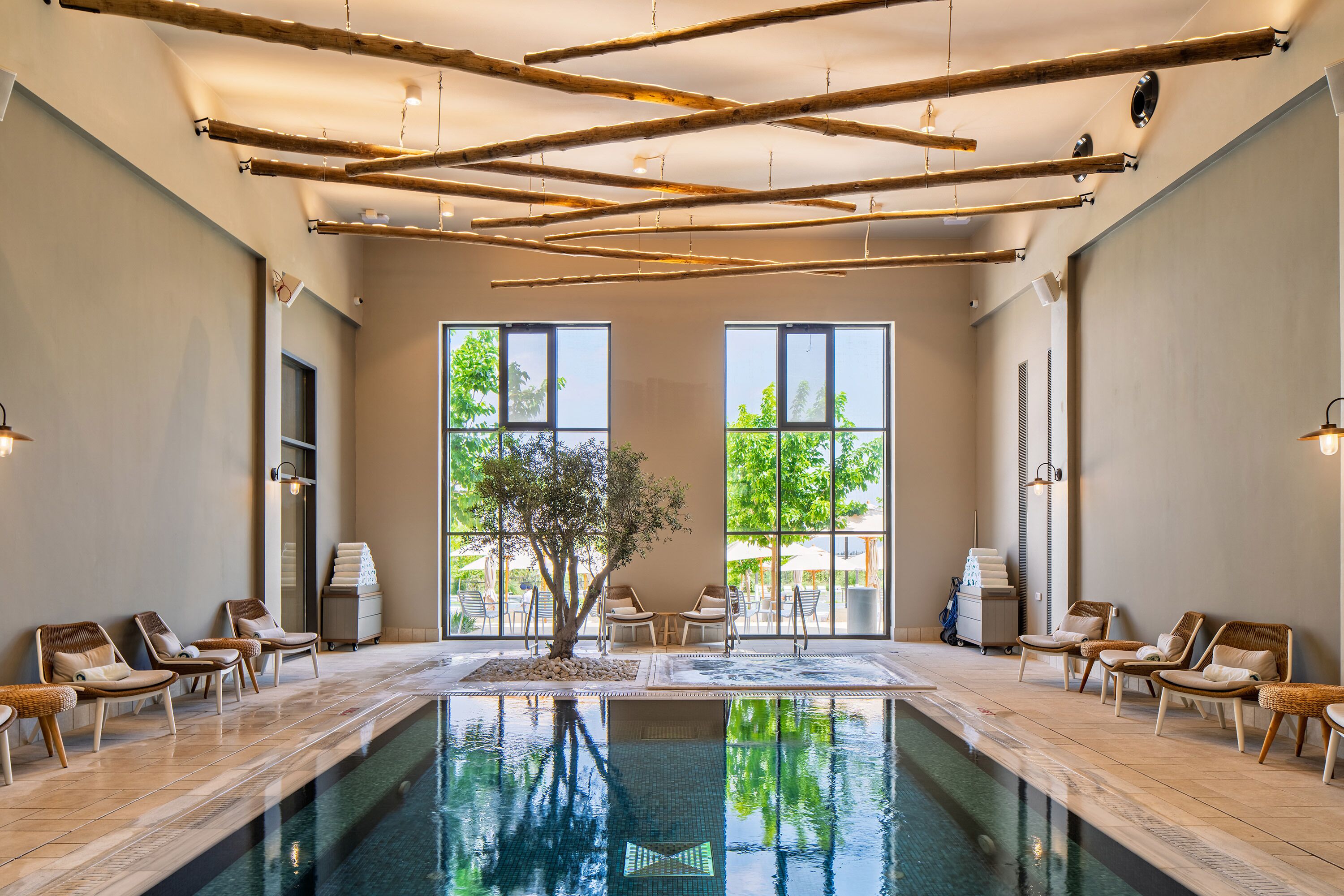 A large indoor swimming pool with lounge chairs and a tree in the center, surrounded by large windows.