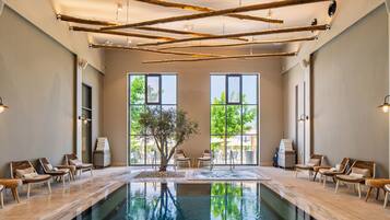 A large indoor swimming pool with lounge chairs and a tree in the center, surrounded by large windows.