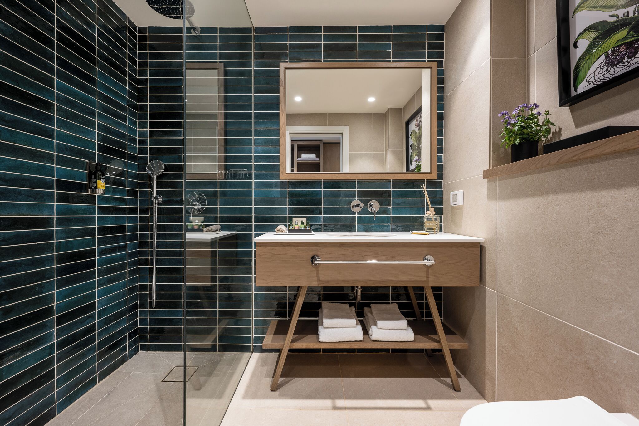 Un baño moderno con una pared de azulejos azul oscuro, un mueble de tocador de madera, un espejo grande y una zona de ducha.