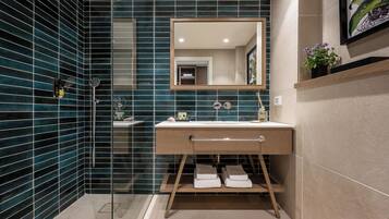 Un baño moderno con una pared de azulejos azul oscuro, un mueble de tocador de madera, un espejo grande y una zona de ducha.