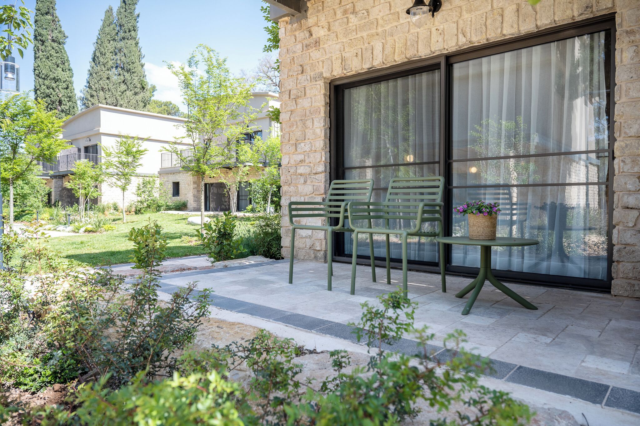 A patio with green chairs and a table, a stone wall, and a large window.