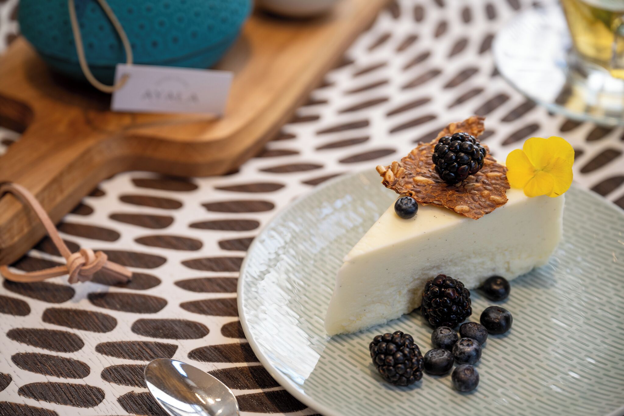 A slice of cheesecake on a plate with blackberries and a yellow flower on top, placed on a table with a patterned tablecloth.