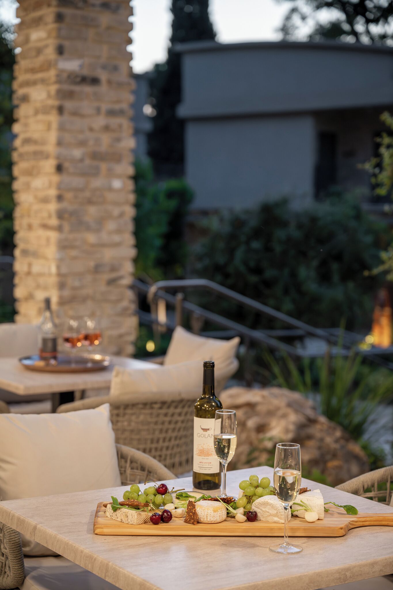 Un espacio exterior para comer con una mesa dispuesta para una comida, destacando una botella de vino, copas, y una variedad de quesos y frutas.