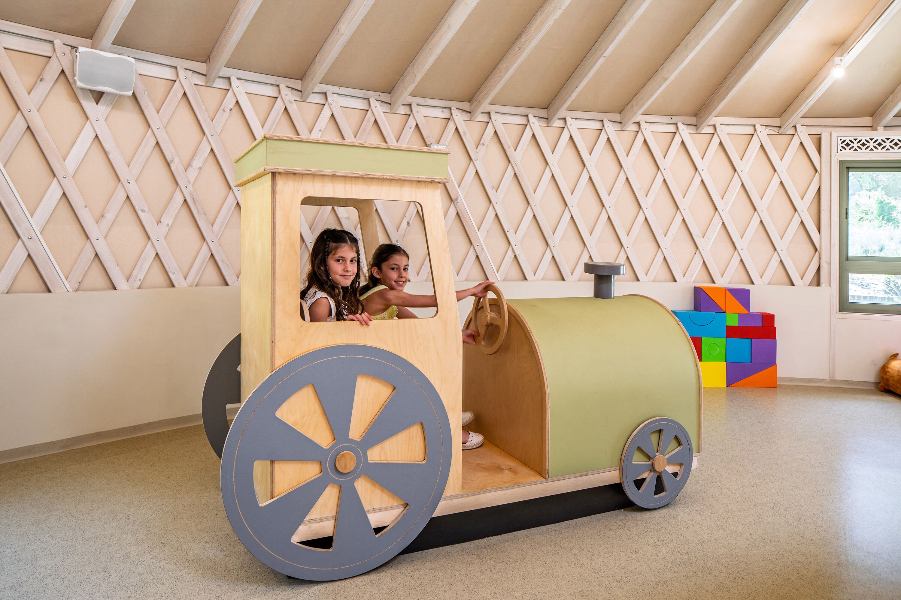 Ein hölzernes Spielhaus mit zwei Kindern darin, einem Fenster und bunten Bauklötzen im Hintergrund.
