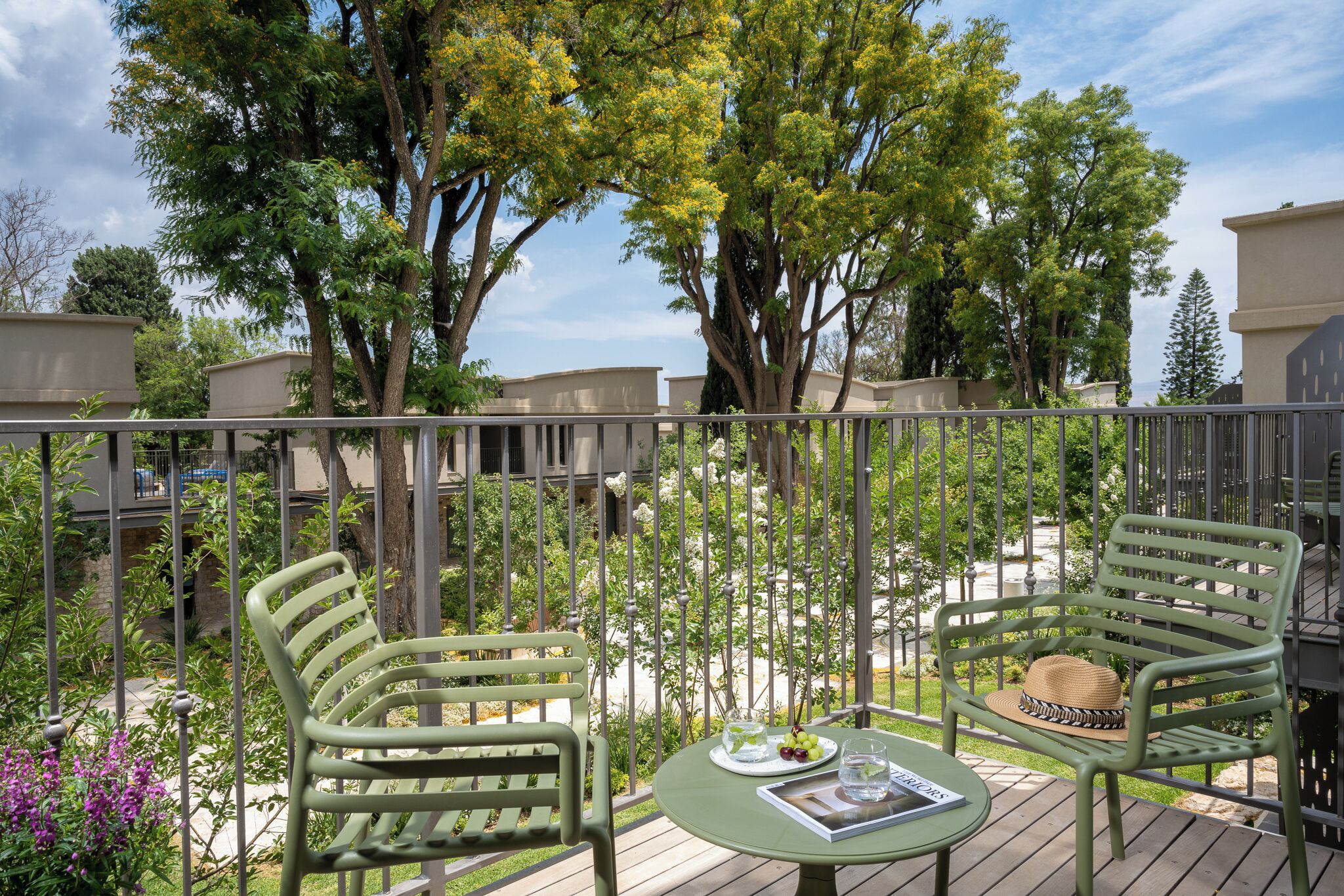 A balcony with two green chairs, a small table with a hat and a plate of fruit, and a view of trees and buildings.
