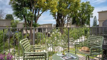 A balcony with two green chairs, a small table with a hat and a plate of fruit, and a view of trees and buildings.