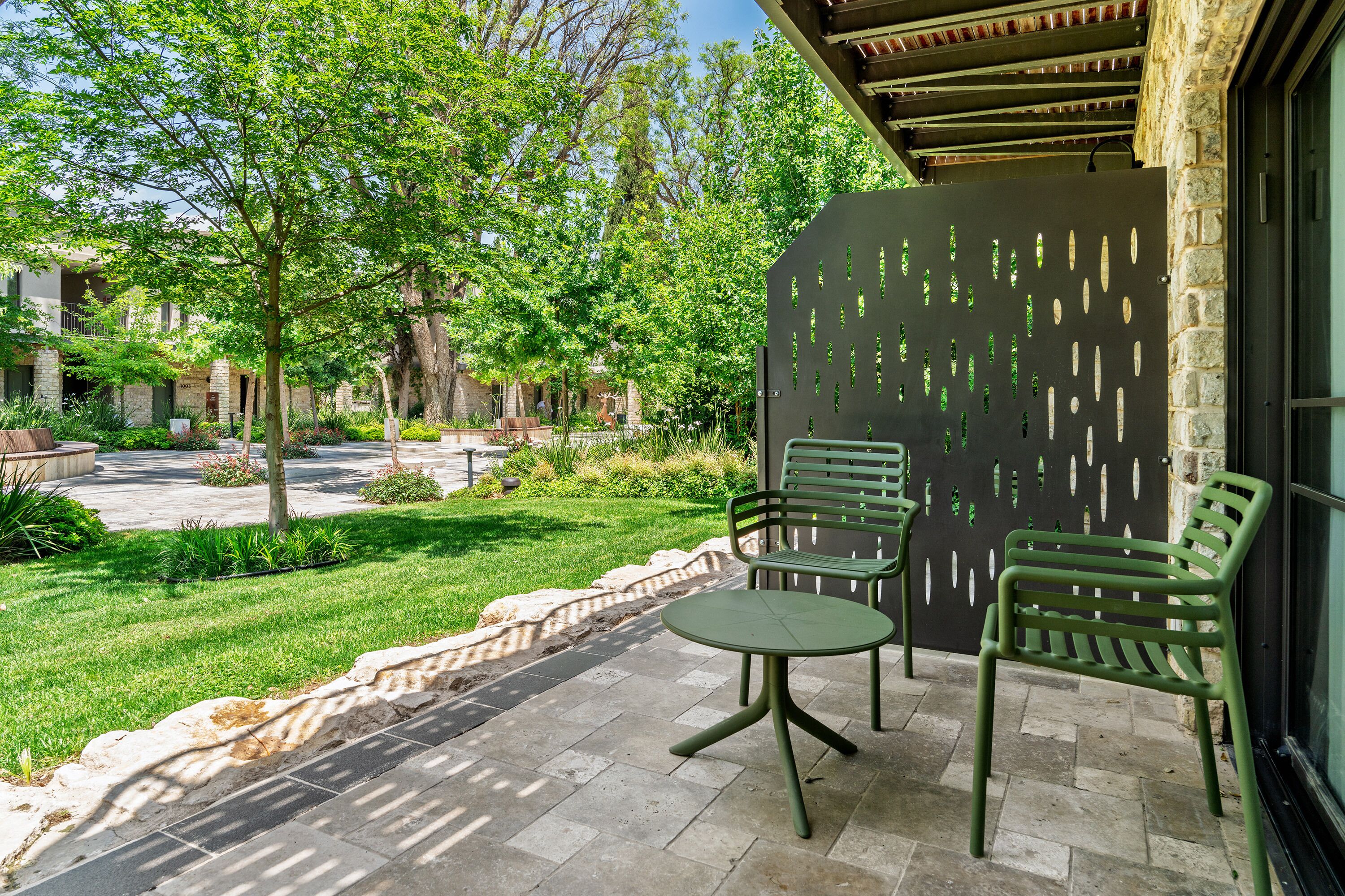Un patio con una mesa y dos sillas, un muro de piedra con un diseño decorativo y vista a un patio interior con árboles y un edificio al fondo.