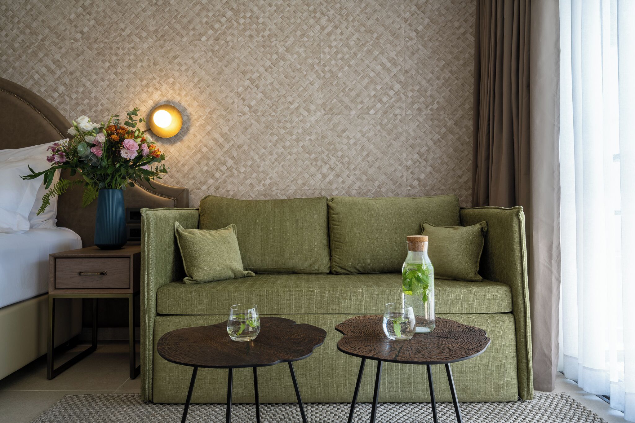 A hotel room with a green sofa, two small round tables, a nightstand with a lamp, and a wall-mounted light fixture.
