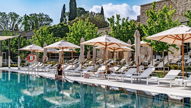 A poolside area with lounge chairs and umbrellas, a clear pool, and a stone building in the background. - Ayala by Isrotel Collection (HaGalil HaElyon)