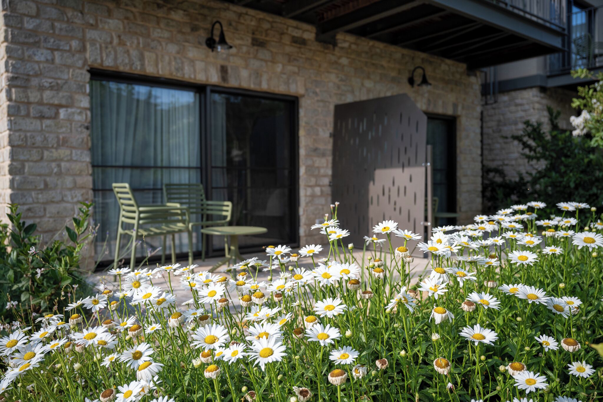 A stone building with a patio area featuring green chairs and a flower bed with white daisies.