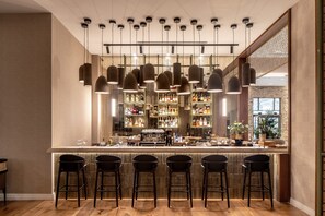 A bar area with multiple hanging pendant lights, a bar counter with stools, and shelves stocked with bottles. - Ayala by Isrotel Collection (HaGalil HaElyon)