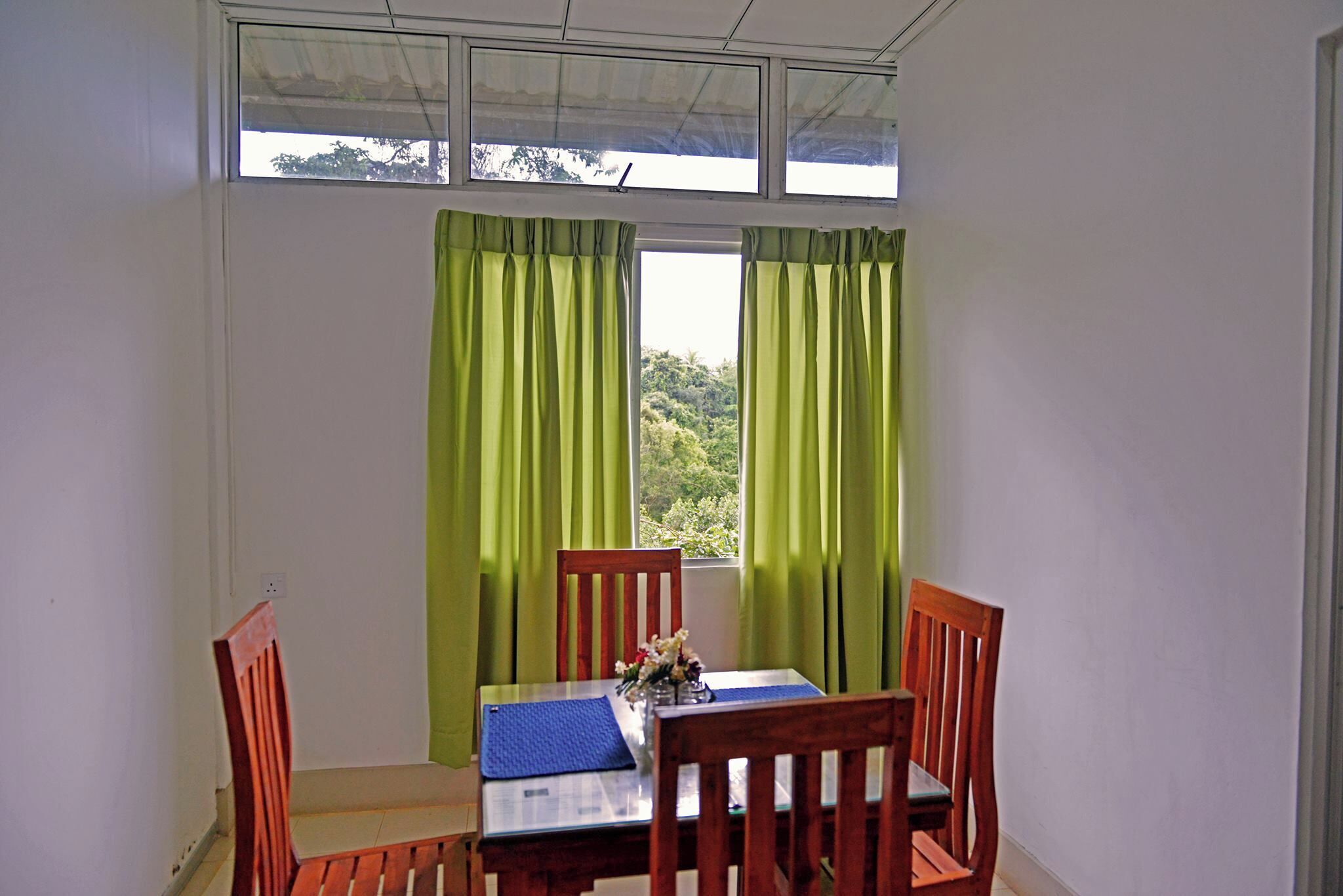 Ein Essbereich mit Tisch und vier Stühlen, ein Fenster mit grünen Vorhängen und Blick ins Grüne.