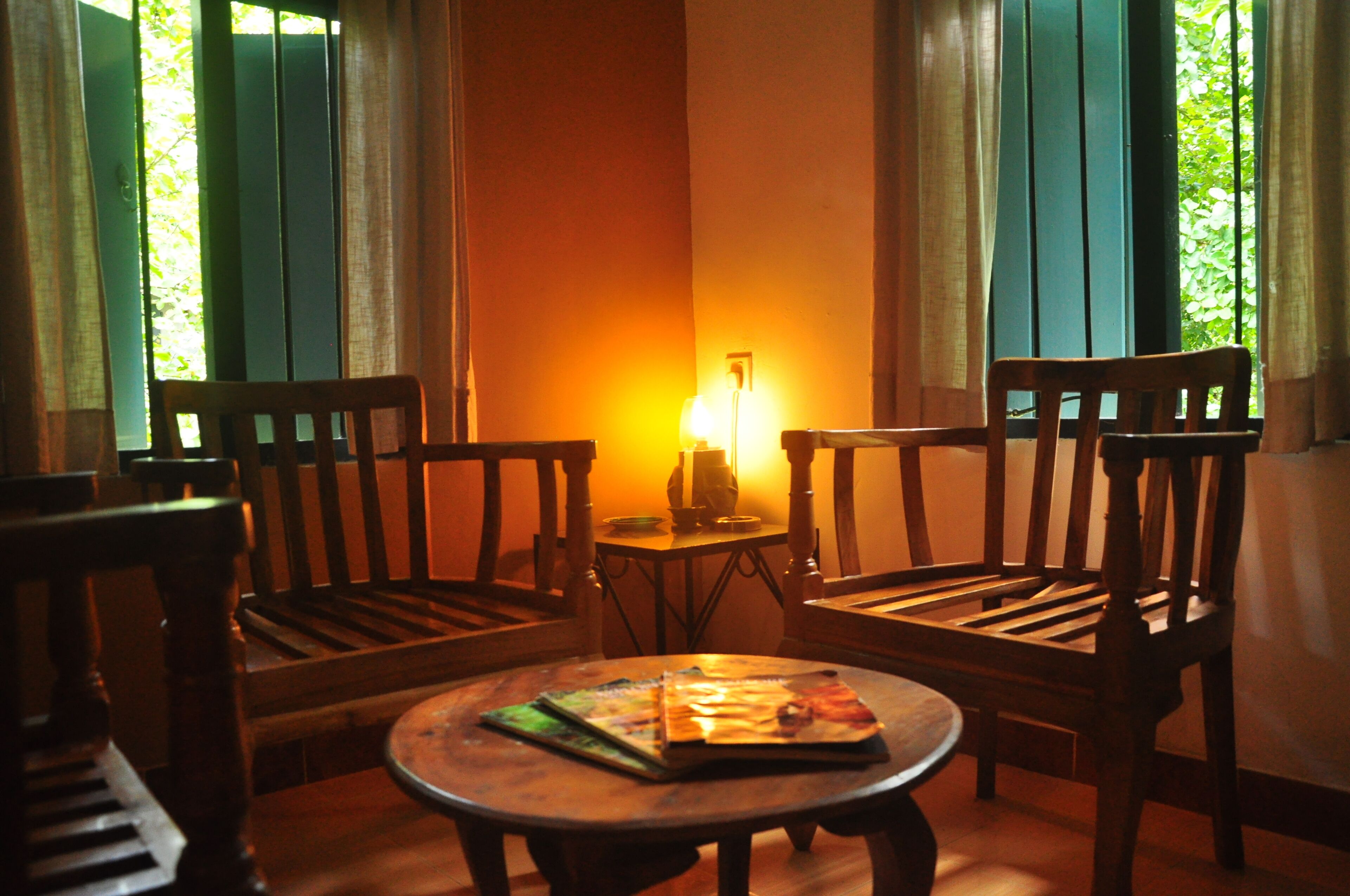 A cozy room with two wooden chairs and a small table, a lamp on a side table, and a view of greenery through the windows.