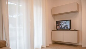 Un salon moderne avec un téléviseur à écran plat posé sur un meuble blanc, des rideaux légers et un canapé beige.