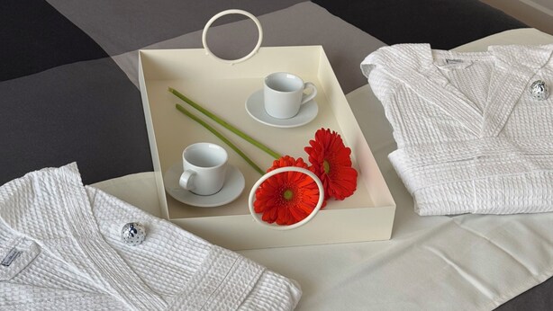 A neatly arranged bed with a white quilt, two white towels, a tray with white cups, green stems, and red flowers.