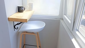 Una habitación pequeña y luminosa con una silla blanca, una estantería de madera con una taza de café y una ventana.