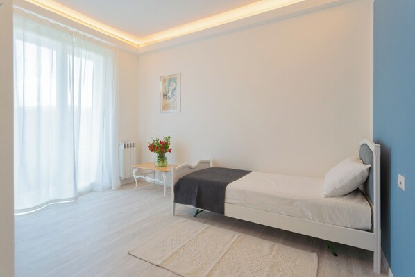A bedroom with a bed, a small table with a vase of flowers, and a window with sheer curtains. - Acqua Chiara Apartment by Rent all Como (Dongo)