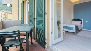 Ein Balkon mit Tisch und Stühlen, ein Bett mit blauer Kopfteil und Blick auf Gebäude im Hintergrund.