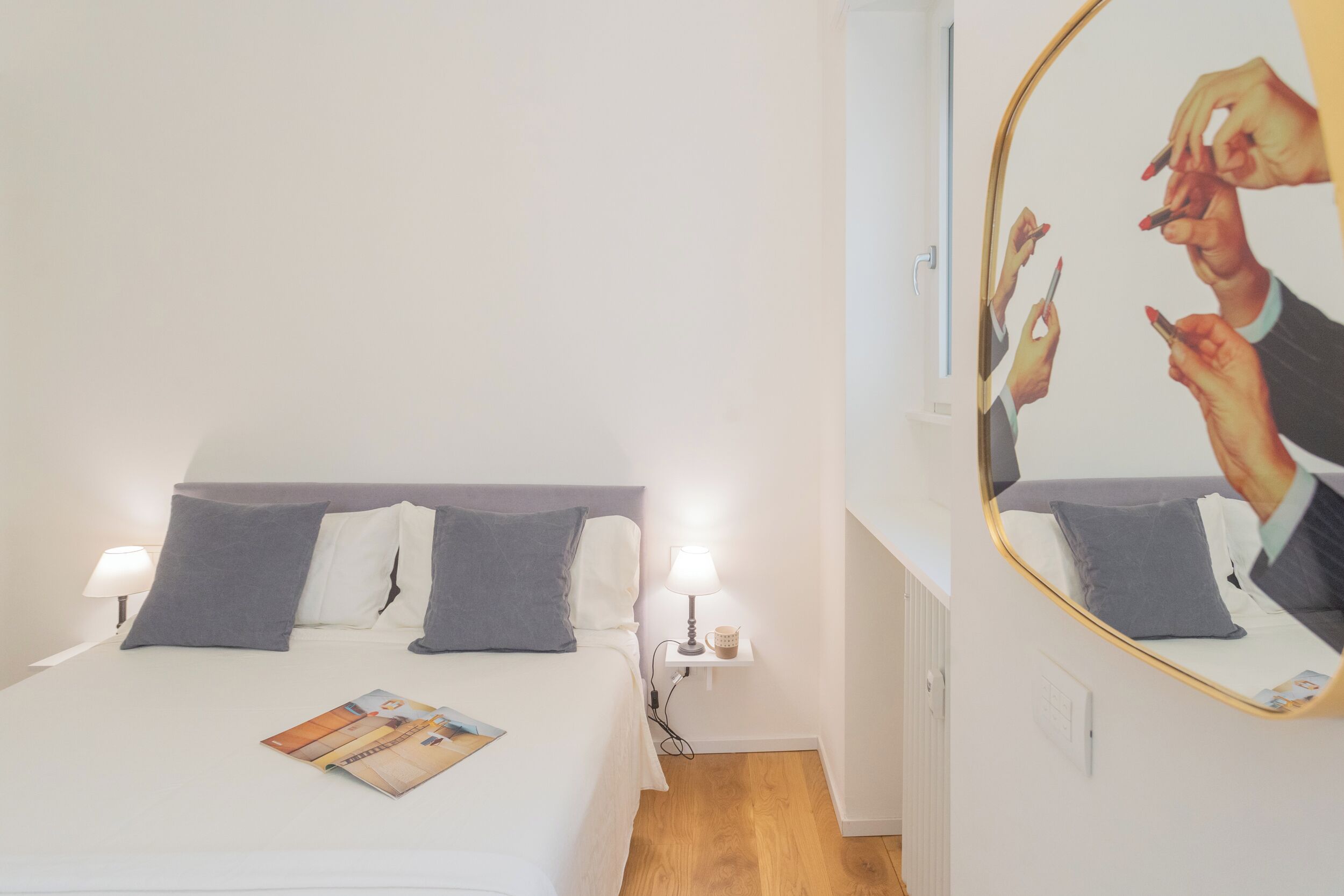 A hotel room with a bed, pillows, a bedside table with a lamp, a mirror, and a magazine on the bed.