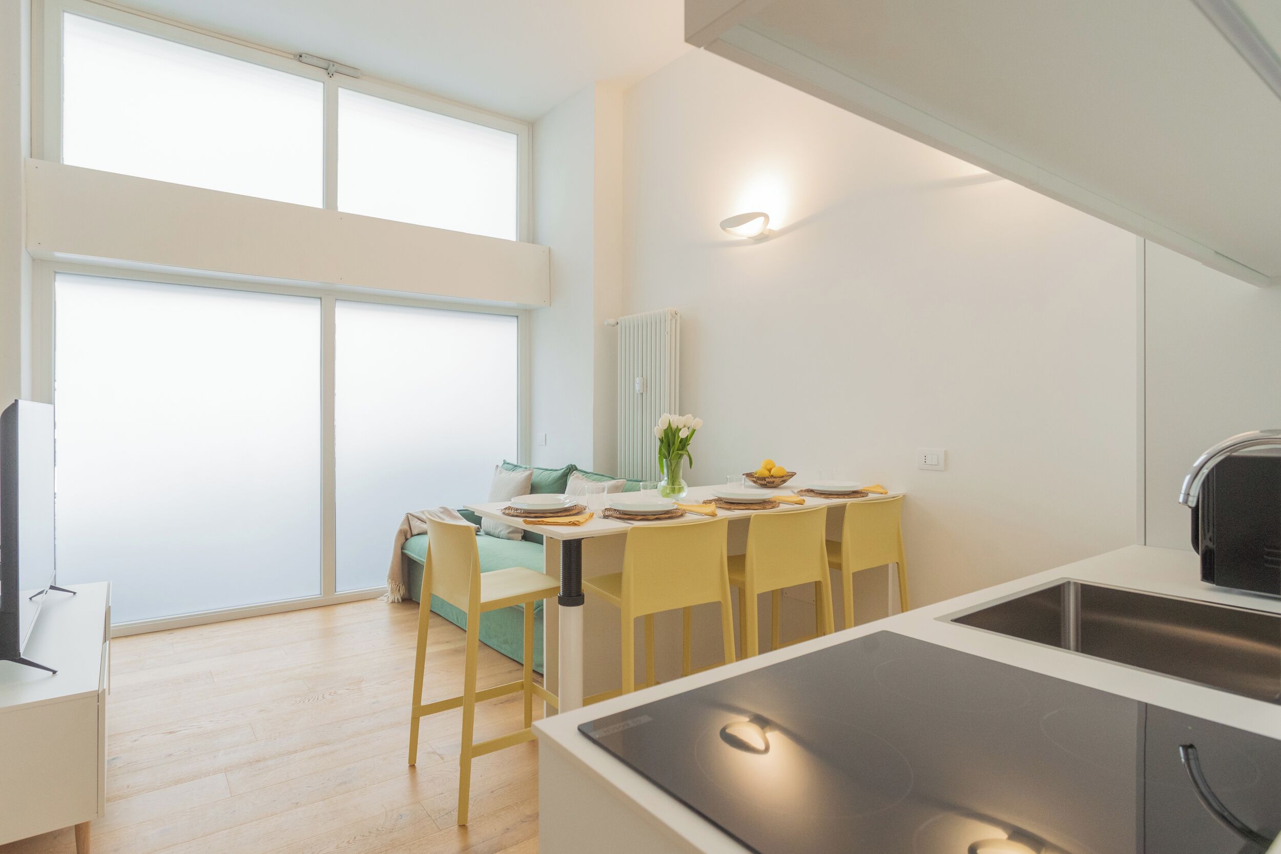 Een moderne keuken met een groot eiland, een eethoek met gele stoelen, en een uitzicht op een stadsgezicht door grote ramen.