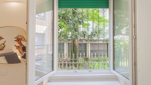 A room with a large window, a green blind, and a view of a tree and fence outside. - A Step From the Lake Apartment by Rent all Como (Como)