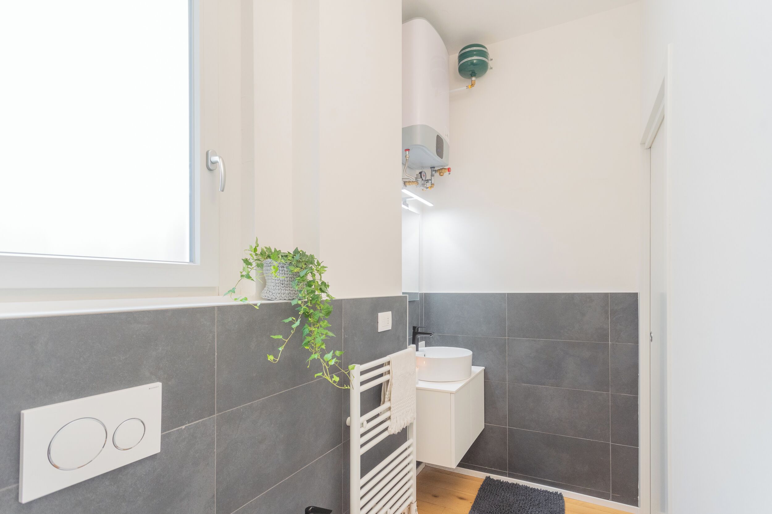 A modern bathroom with grey tiles, a white sink, a towel rack, and a window.