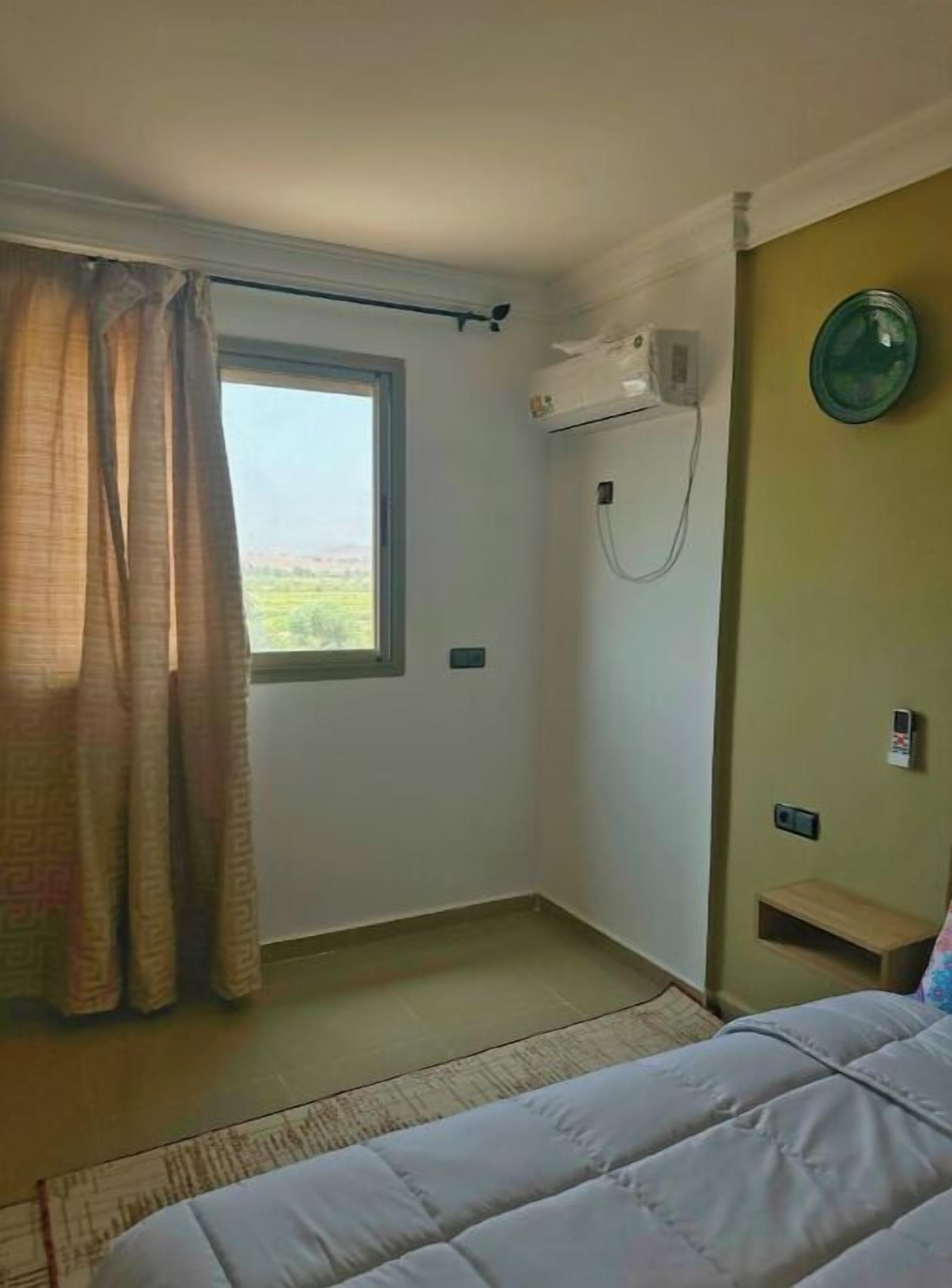 Una habitación pequeña y poco amueblada con una cama individual, una ventana con cortinas, un equipo de aire acondicionado y un estante pequeño.