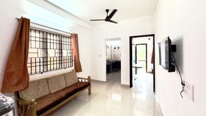 A spacious room with a wooden bench, a ceiling fan, a TV mounted on the wall, and a window with metal bars and curtains. - Sishya Service Apartment - Chennai OMR  (Chennai)