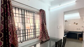 A dining area with a glass table and chairs, a window with metal bars, and a kitchen visible in the background. - Sishya Service Apartment - Chennai OMR  (Chennai)