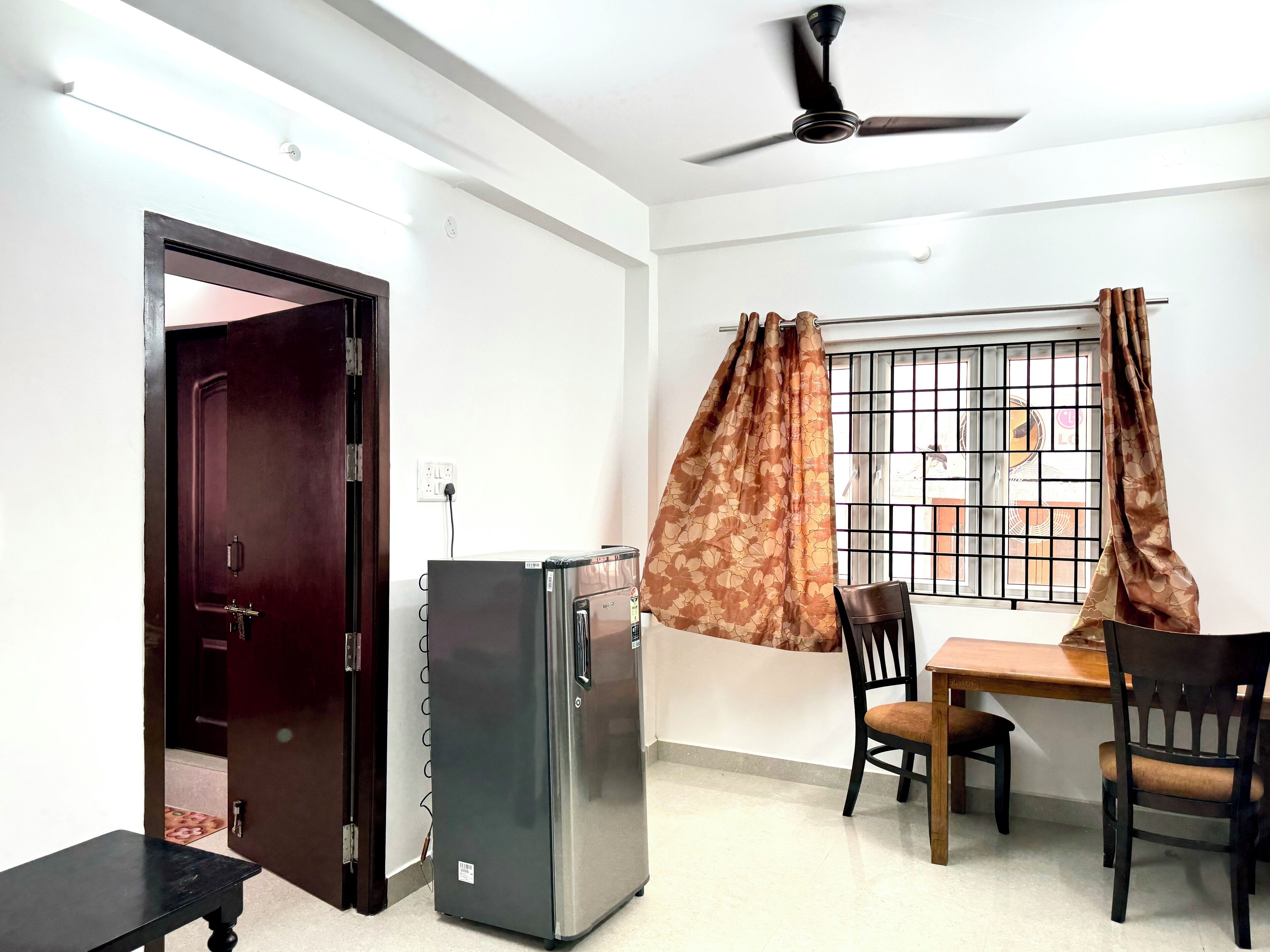 A room with a dining table, two chairs, a ceiling fan, a refrigerator, and a window with metal bars and curtains.