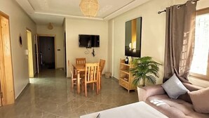 A hotel room with a bed, a TV, a dining table with chairs, a plant, and a window with curtains.