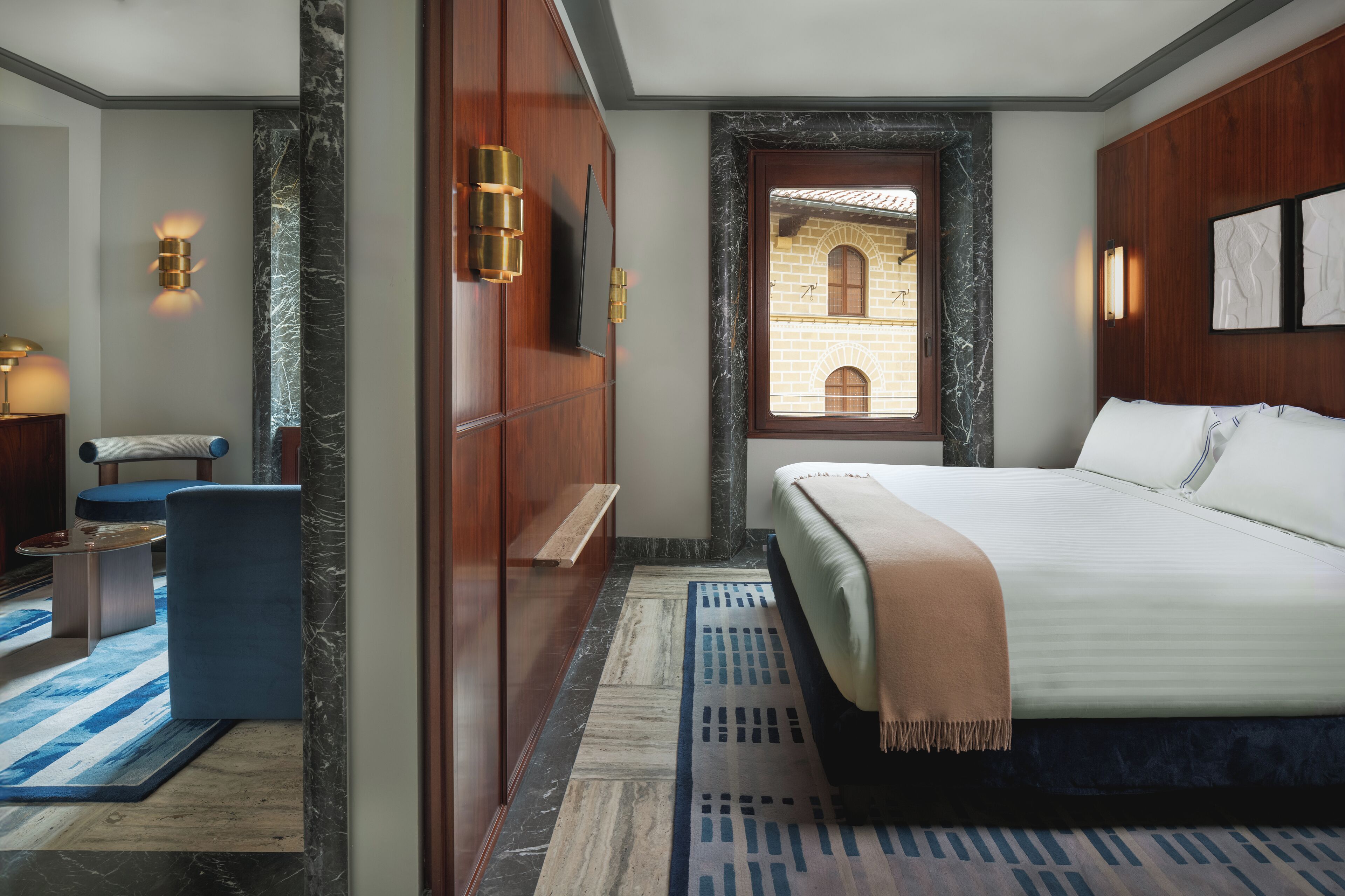 A hotel room with a large bed, a seating area with a chair and table, a wooden wardrobe, and a view of an outdoor courtyard through a window.
