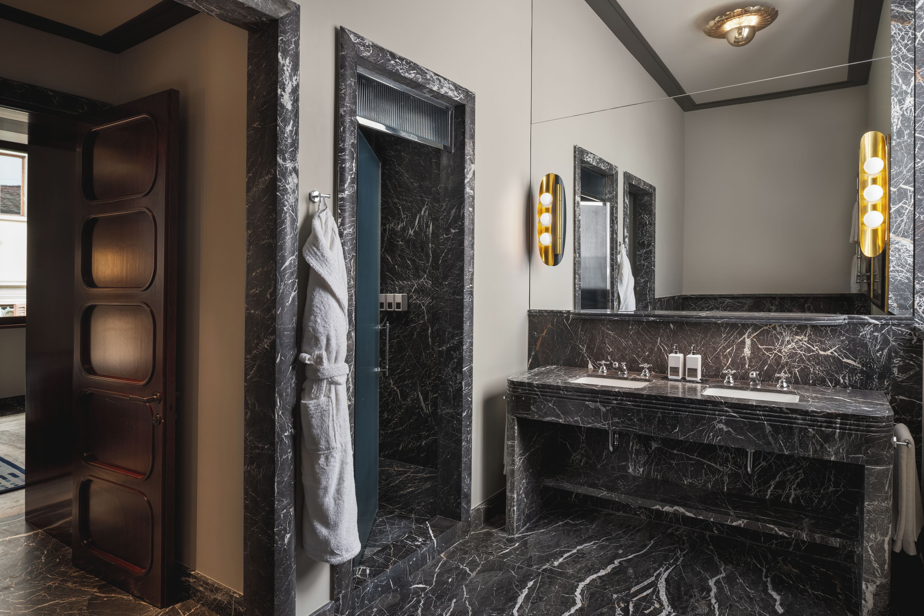 A modern bathroom with a dark marble vanity, a large mirror with lights, and a robe hanging on the door.