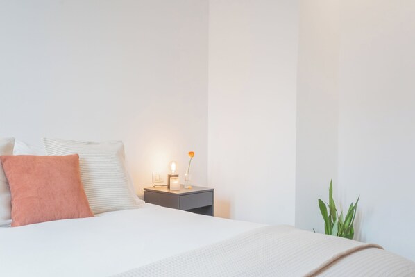Ein ordentlich bezogenes Bett mit weißen und pfirsichfarbenen Kissen, ein Nachttisch mit Lampe und ein Blumentopf.