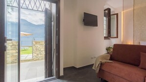 Una habitación con puerta corrediza de vidrio que da a un balcón con vistas a montañas y un lago. Un televisor de pantalla plana está montado en la pared, y un sofá rojo se encuentra apoyado contra ella.