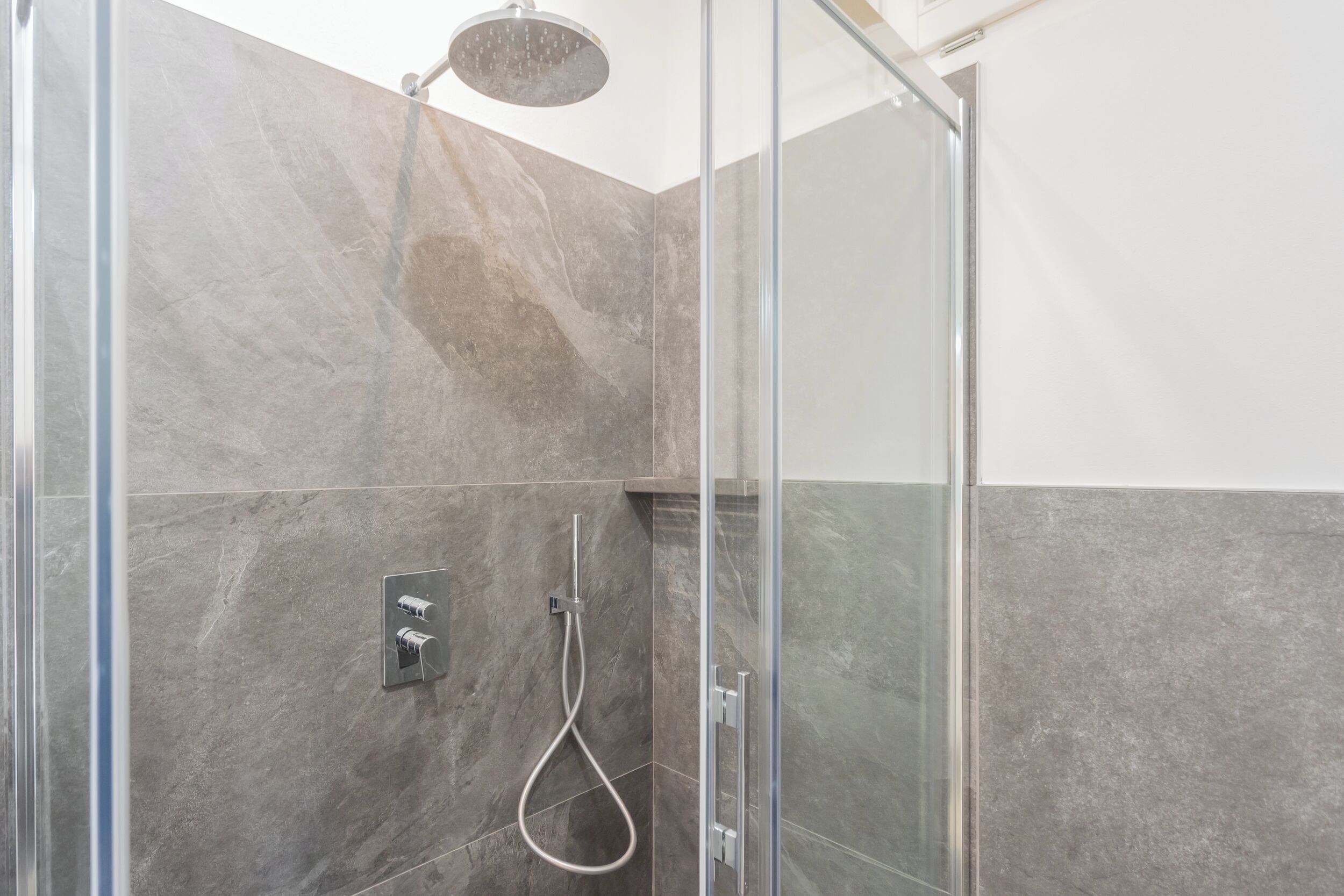 Une douche moderne dotée d’un pare-douche en verre, d’un pommeau de douche à main et d’un panneau de commande fixé au mur.