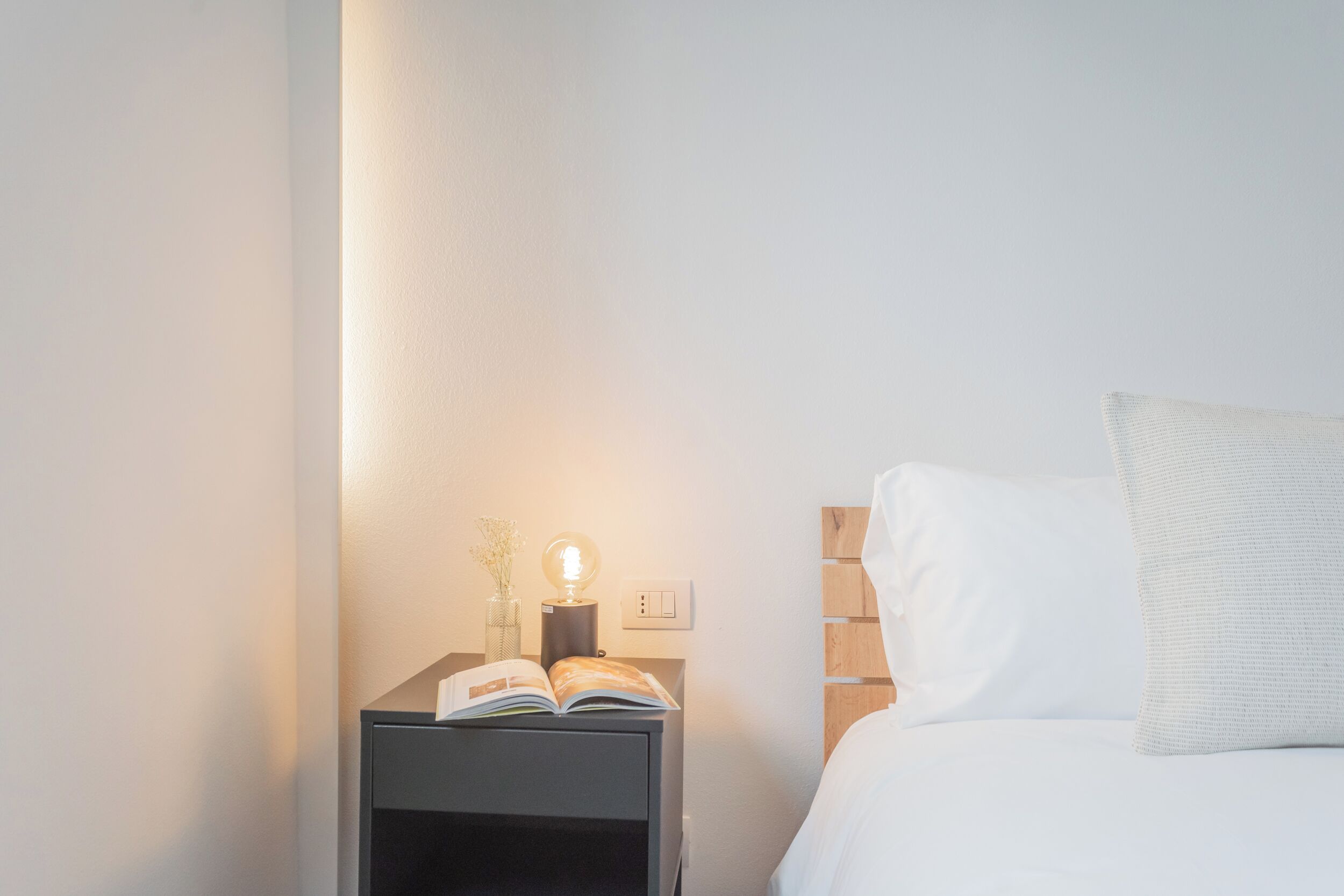 Un lit bien fait, avec une literie blanche et un oreiller, une table de chevet avec un livre et une lampe, et un luminaire fixé au mur.